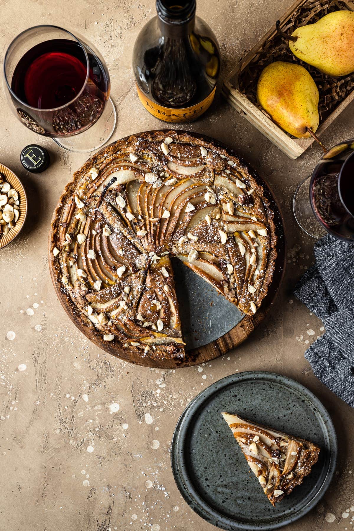 A golden brown pear tart with a slice cut out and resting on a blue grey ceramic plate nearby. Surrounding the tart are a bowl of chopped hazelnuts, pears in a wooden crate, a wine bottle and two wine glasses filled with red wine, and a dark blue linen napkin. Everything rests on a textured tan background.