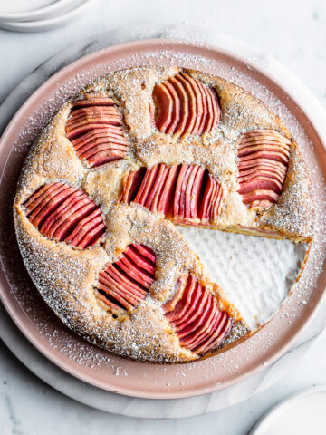 A round cake dusted with powdered sugar has a slice cut out of it. It sits on a pink plate atop a white marble platter, which rests on a white marble surface. There are white plates at the top and bottom of the frame.