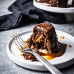 Close up of a mini cake on a white plate cut in half, revealing a moist crumb with bits of apples inside. The cake is covered in a drizzle of salted caramel sauce. There are other cakes on a platter in the background with a navy blue napkin.