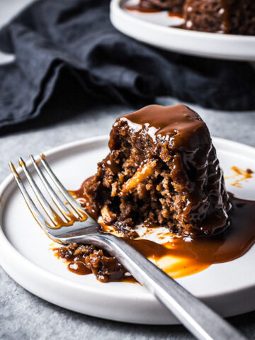 Close up of a mini cake on a white plate cut in half, revealing a moist crumb with bits of apples inside. The cake is covered in a drizzle of salted caramel sauce. There are other cakes on a platter in the background with a navy blue napkin.