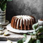 Brown bundt cake with white glaze on a marble platter with apple branches in foreground