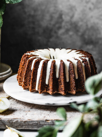 Brown bundt cake with white glaze on a marble platter with apple branches in foreground