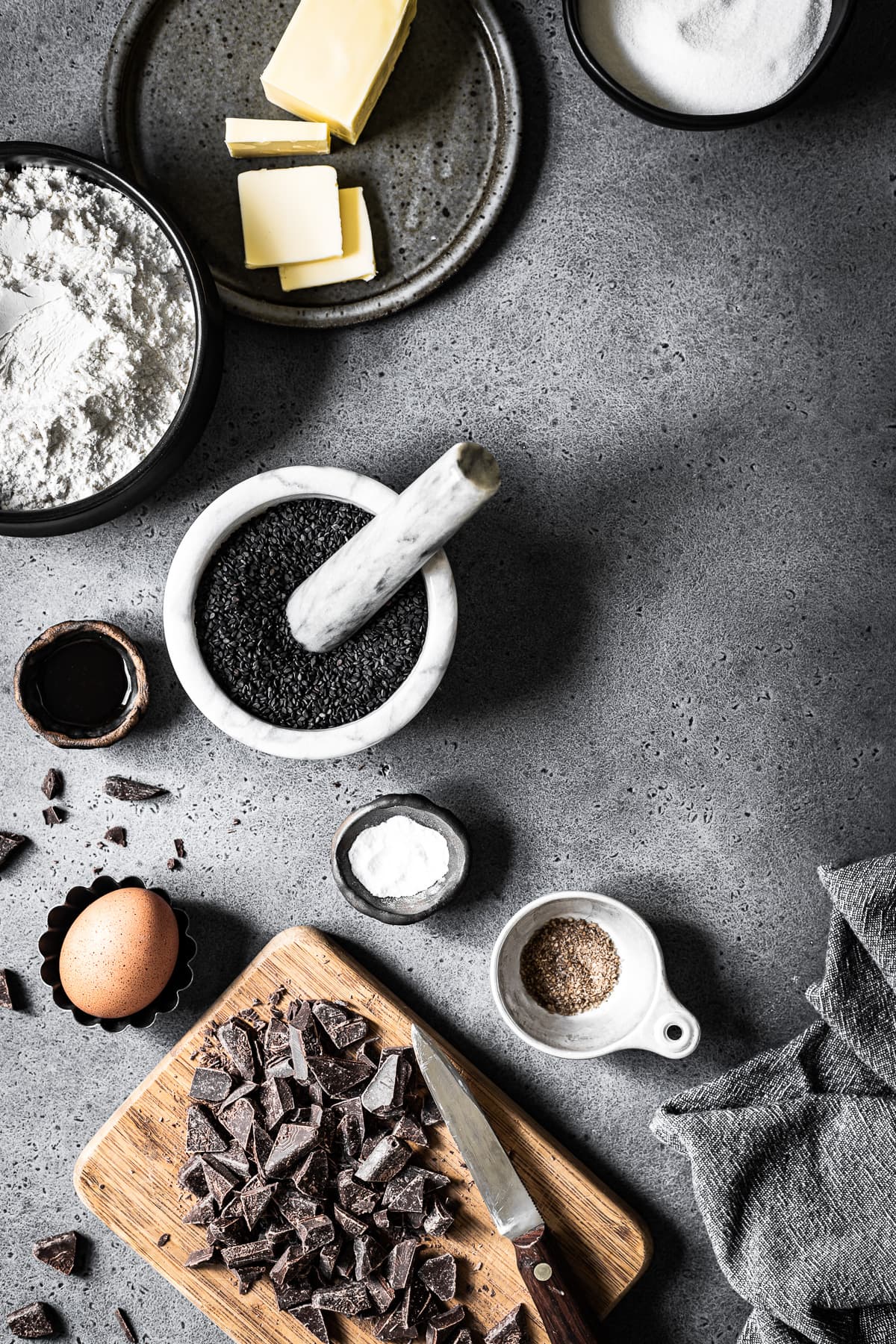 Various bowls and containers filled with ingredients for making cookies arranged on a grey stone background. The light is entering from left. The image is well lit and there are dark shadows.