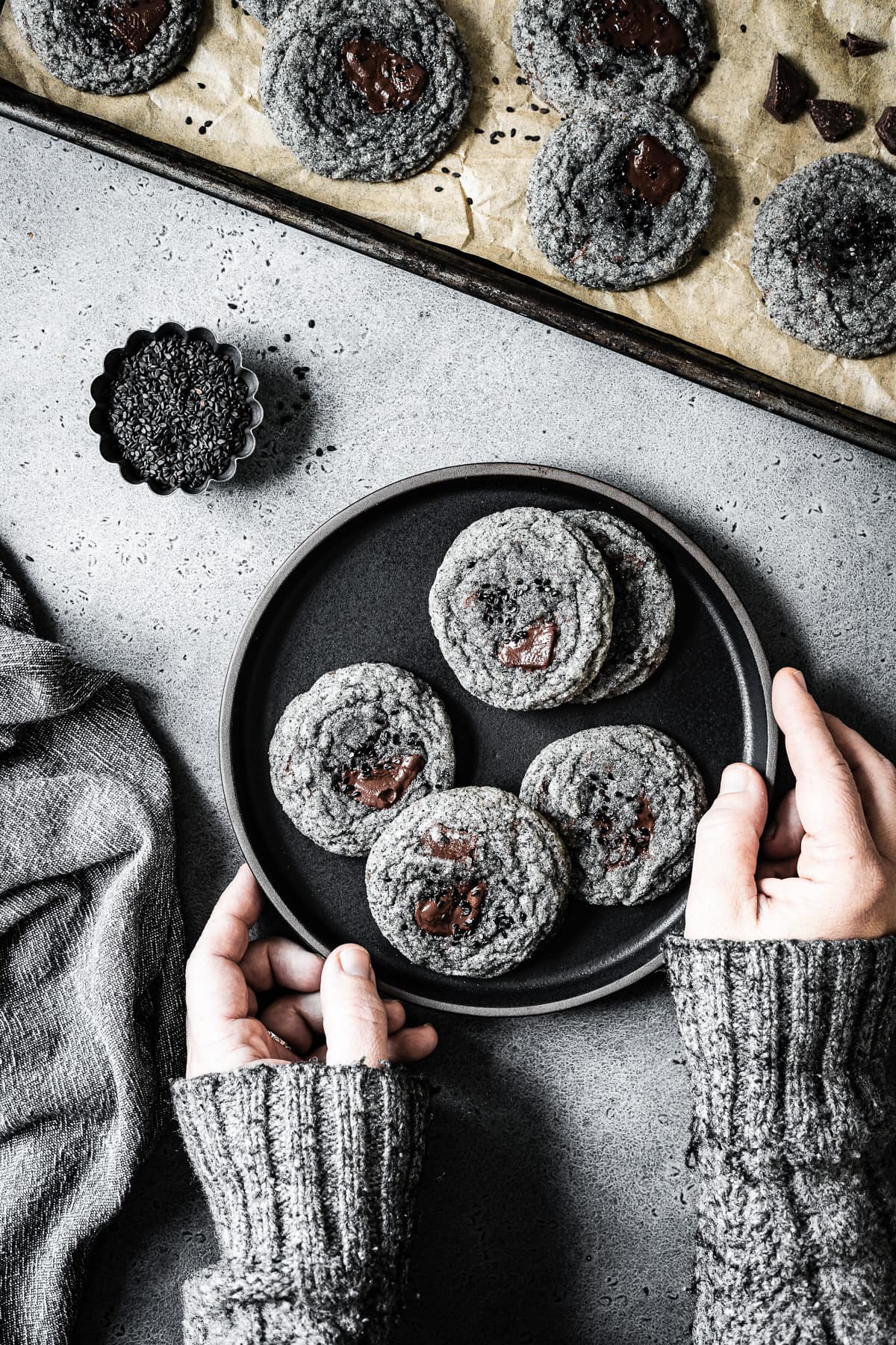 Arms in a grey sweater reach into the frame to hold a black plate of cookies. The colors in the image are monochromatic: grey, black and brown. The sweater color and texture matches the grey stone background and the color of the cookies.
