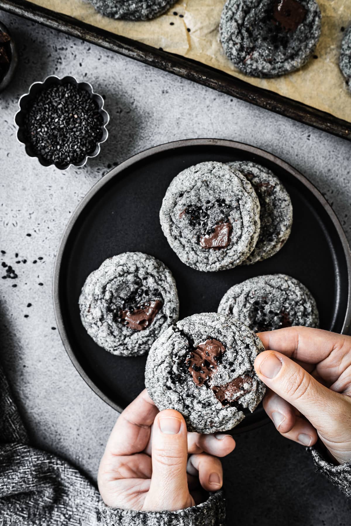 Hands hold a cookie which is being broken in half. There is a black plate full of cookies in the background on a grey stone surface. A small container of black sesame seeds sits nearby.