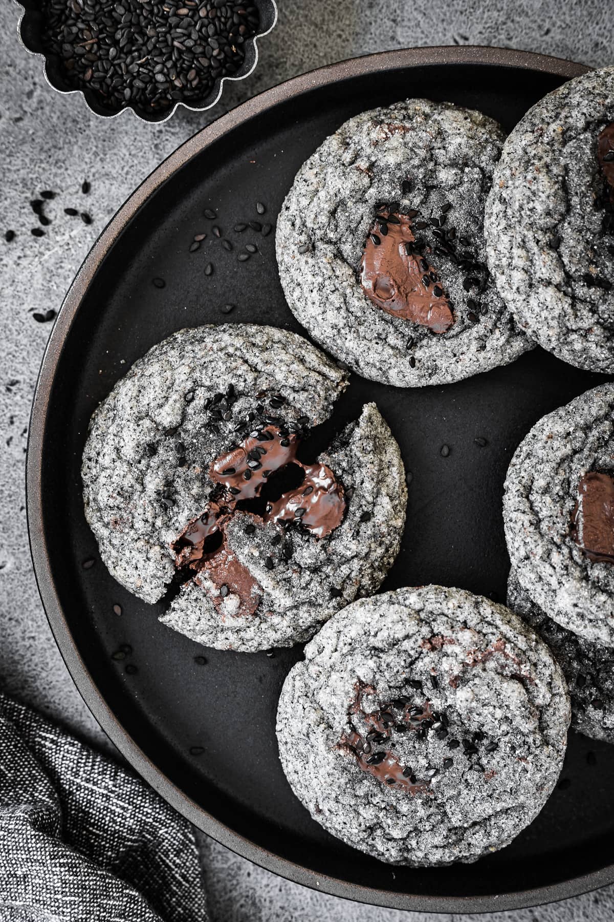 A closeup photo of cookies on a black plate, resting on a grey stone surface. The cookies have pools of melted chocolate and are sprinkled with black sesame seeds.