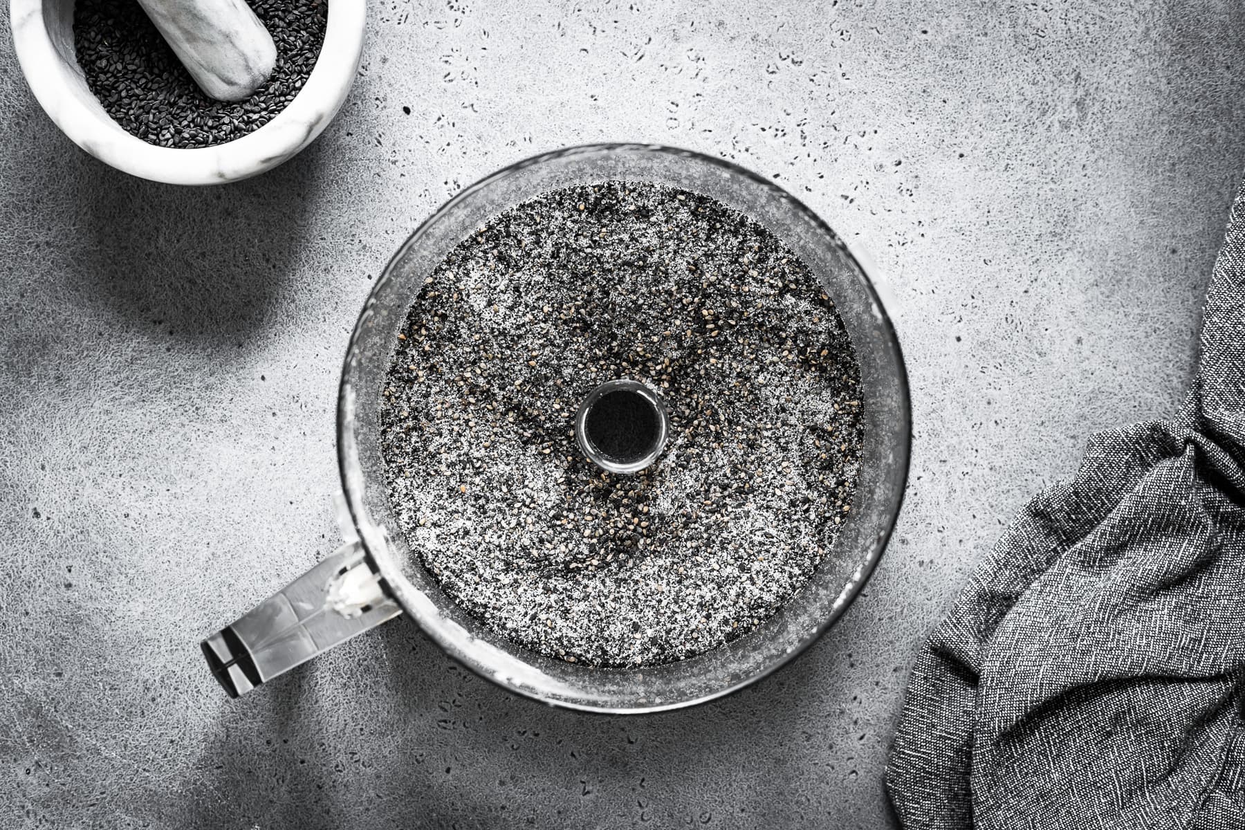 Recipe process shot showing sugar and black sesame seeds ground in a food processor on a grey stone surface.