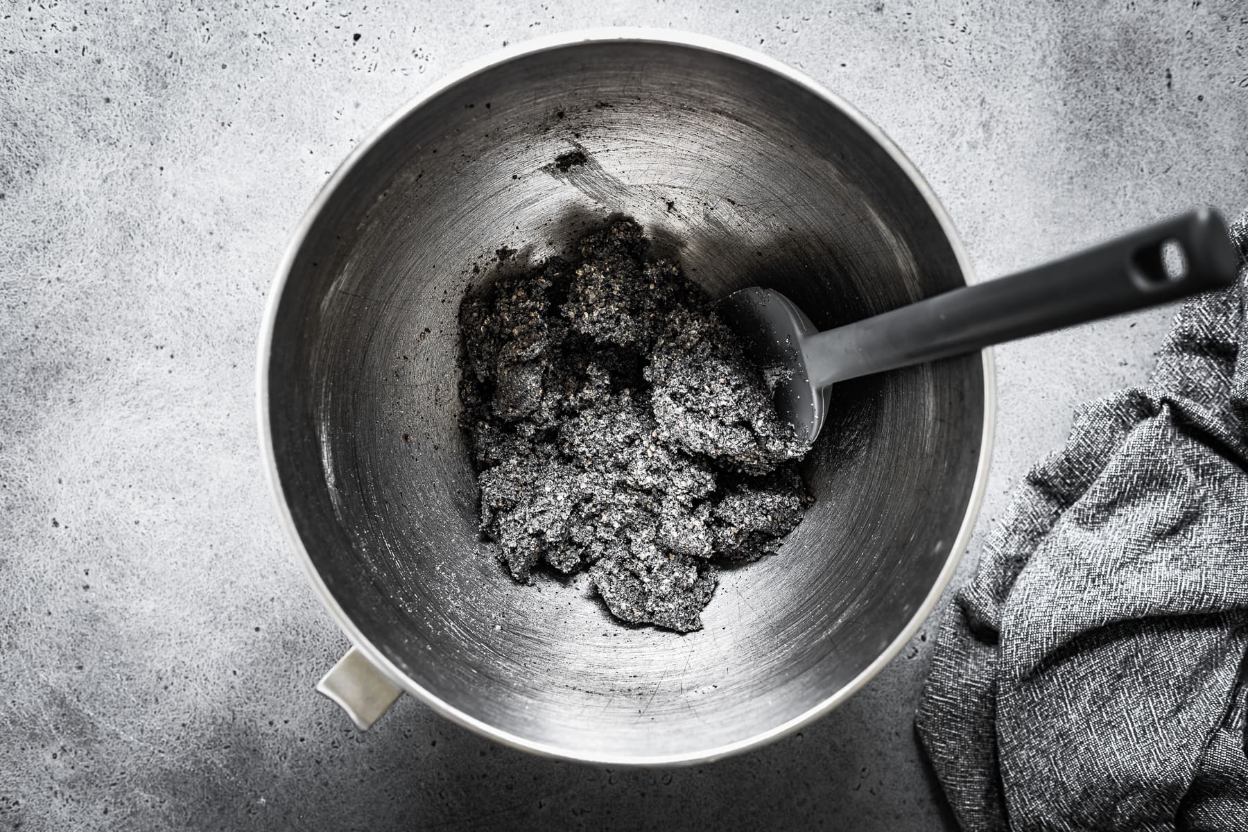 Recipe process shot showing cookie dough with butter, sugar and sesame seeds added. The silver mixing bowl sits on a grey stone surface. There is a grey spatula in the bowl.