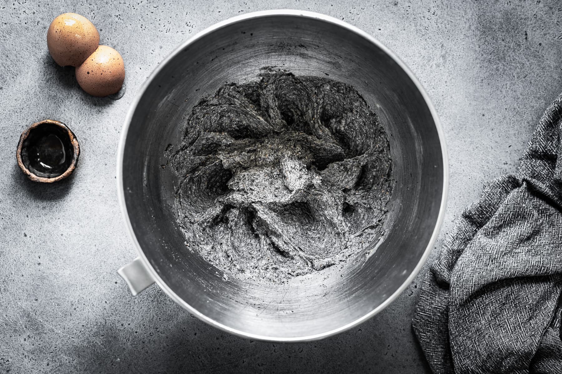 Recipe process shot showing cookie dough with eggs and vanilla added. The silver mixing bowl sits on a grey stone surface.