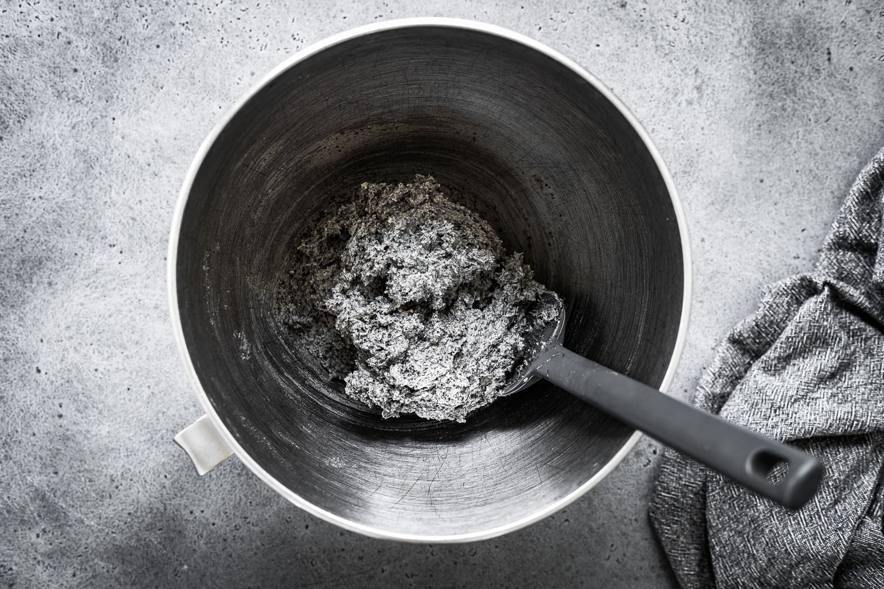 Recipe process shot showing cookie dough in a silver mixing bowl with a grey spatula. The bowl rests on a grey stone surface.