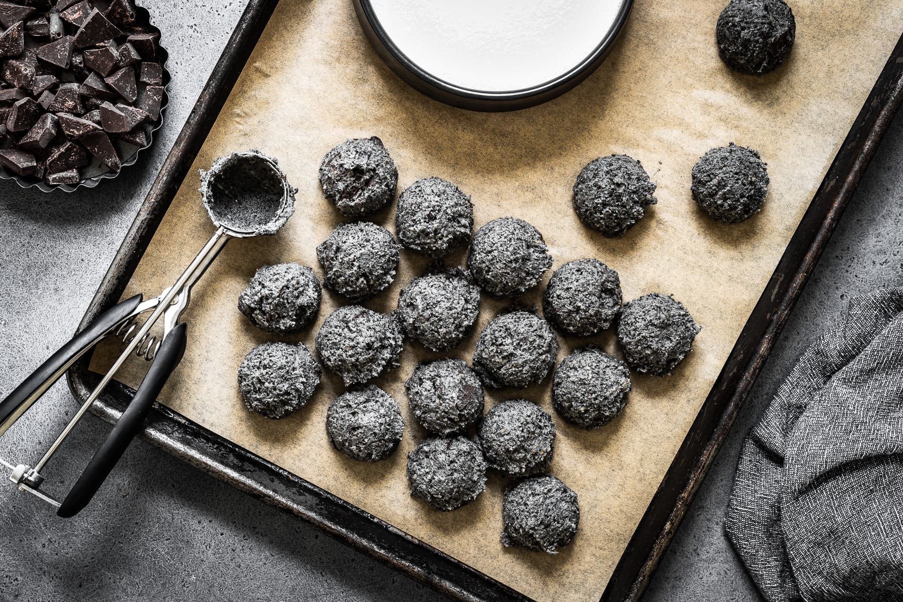 Scoops of cookie dough on a tan parchment lined baking sheet with the cookie scoop at left. A bowl of granulated sugar is at top. At left is a container of chocolate chunks. The pan sits on a grey stone background.