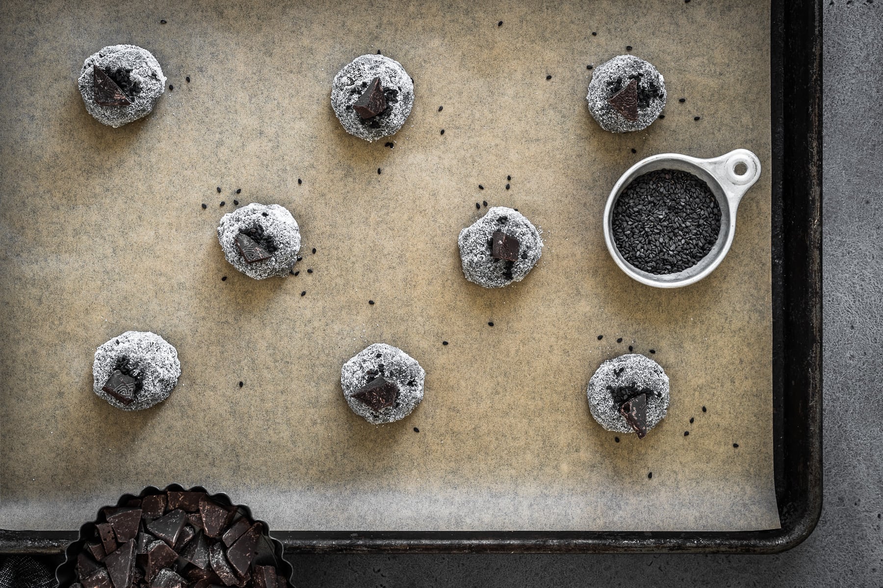 Balls of cookie dough rolled in sugar, sprinkled with seeds and topped with chocolate chunks on a parchment lined baking sheet.