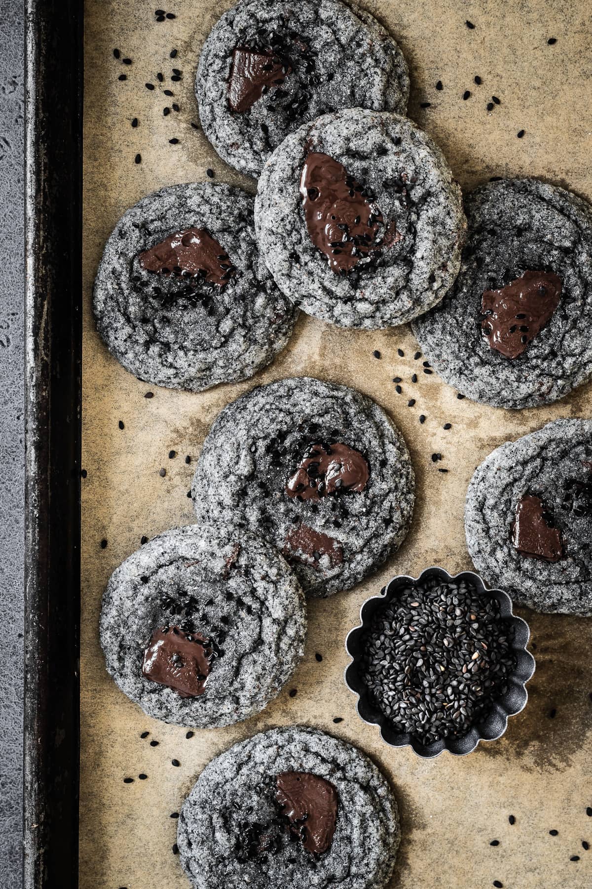 Grey cookies with pools of melted chocolate and sesame seeds on a parchment lined baking sheet.