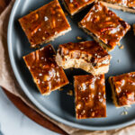 Golden brown squares of caramel pecan bars rest on a blue plate with brown parchment underneath. One of the bars has a bite taken out of it.