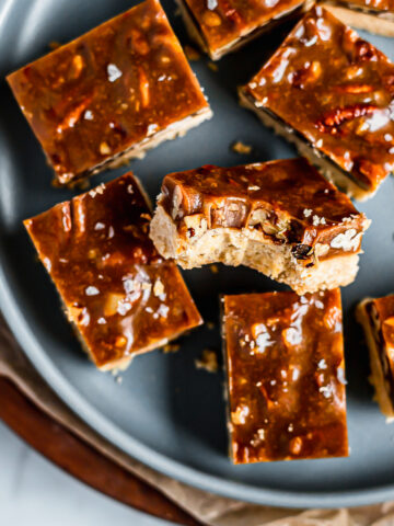 Golden brown squares of caramel pecan bars rest on a blue plate with brown parchment underneath. One of the bars has a bite taken out of it.