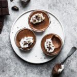 Three chocolate desserts topped with whipped cream rest on a round white plate. There is a bite missing from one of the containers. A spoon and some chopped chocolate rest nearby on a grey stone surface.