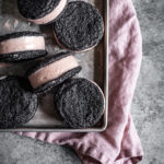 Chocolate cookies sandwich pink hued ice cream. There are six ice cream sandwiches on a vintage metal baking sheet. Tucked under the baking sheet is a pink linen napkin. It all rests on a grey stone surface.