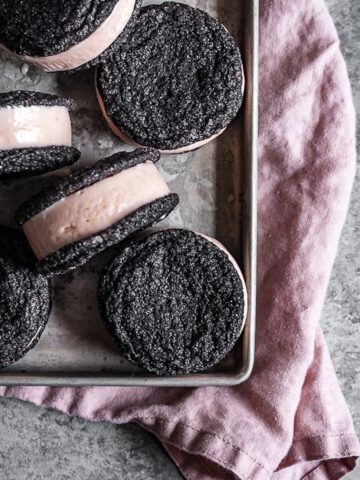 Chocolate cookies sandwich pink hued ice cream. There are six ice cream sandwiches on a vintage metal baking sheet. Tucked under the baking sheet is a pink linen napkin. It all rests on a grey stone surface.