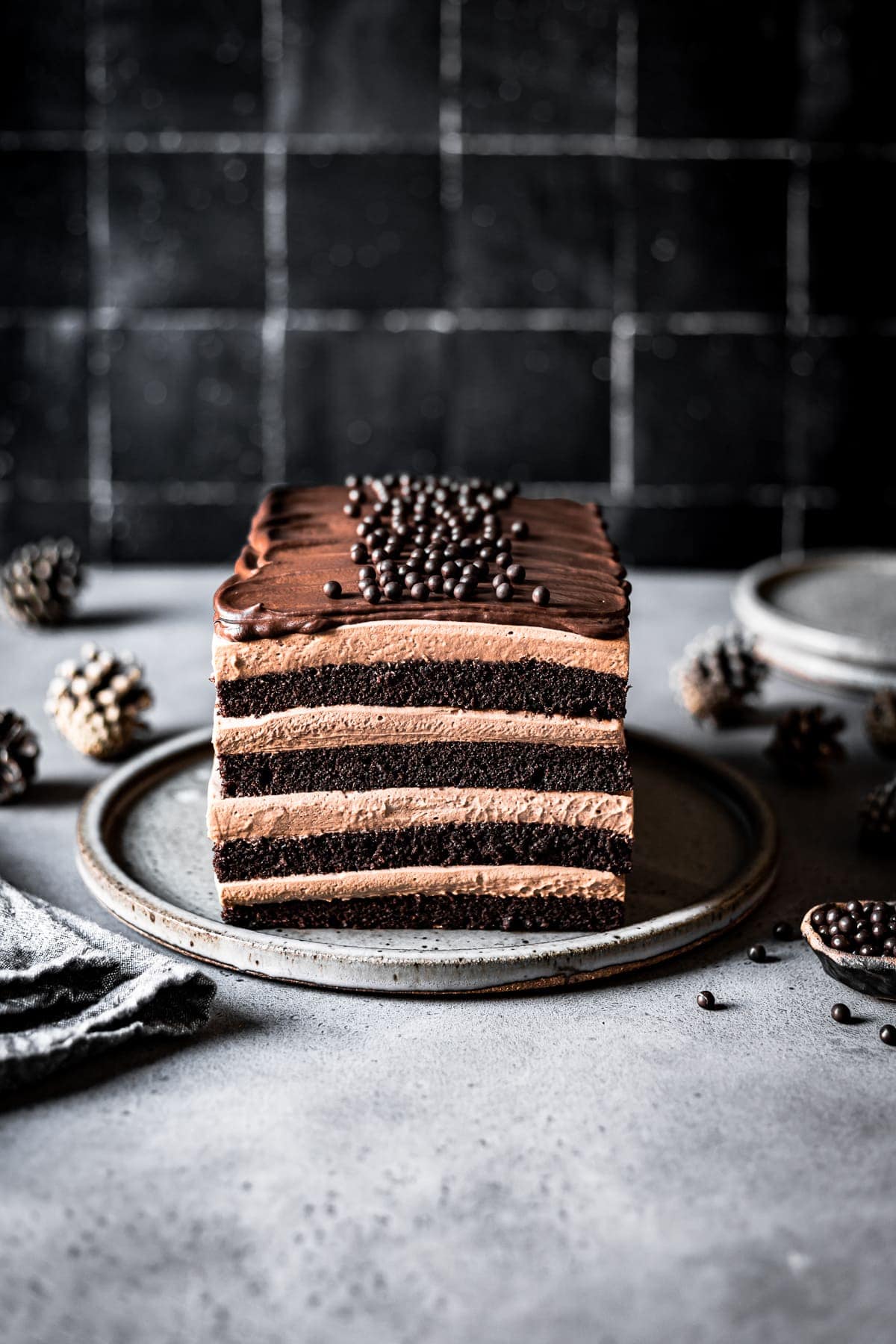 A layered rectangular chocolate cake with mousse filling on a grey plate and grey stone surface with a black tile background. Edible chocolate pearls in a small bowl and two ceramic plates peek out from the right hand of the scene. There are pine cones out of focus in the back of the image. 