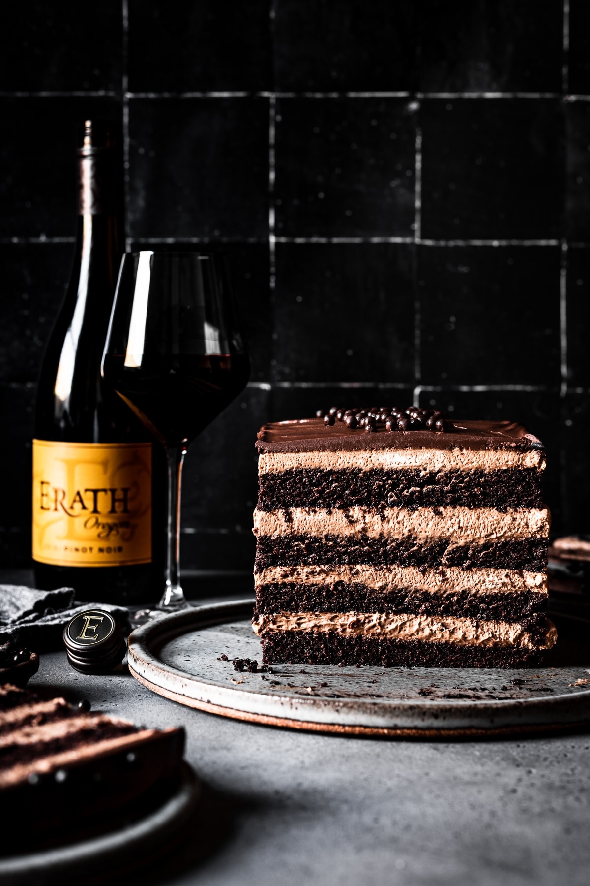 A layered rectangular cake with mousse filling on a grey speckled ceramic plate and grey stone surface with a black tile background. A ceramic plate with a slice of cake peeks out from the left front corner of the scene. A bottle of red wine with an orange-yellow label and a full glass of red wine are slightly out of focus on the left side of the scene. 