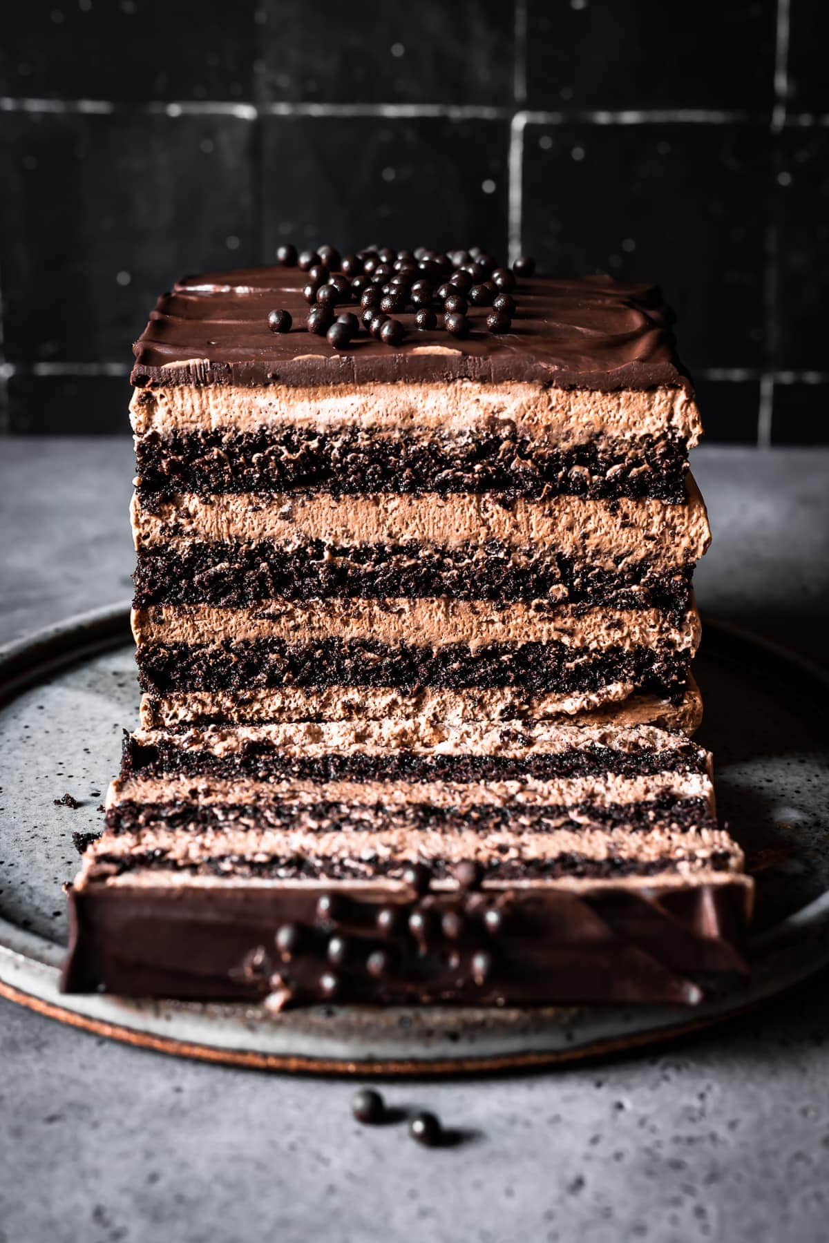 A moody front view image of a slice of chocolate mousse cake resting on its side on a grey speckled ceramic serving platter next to the remainder of the cake. The platter is on a grey stone surface with black tile in the background. 