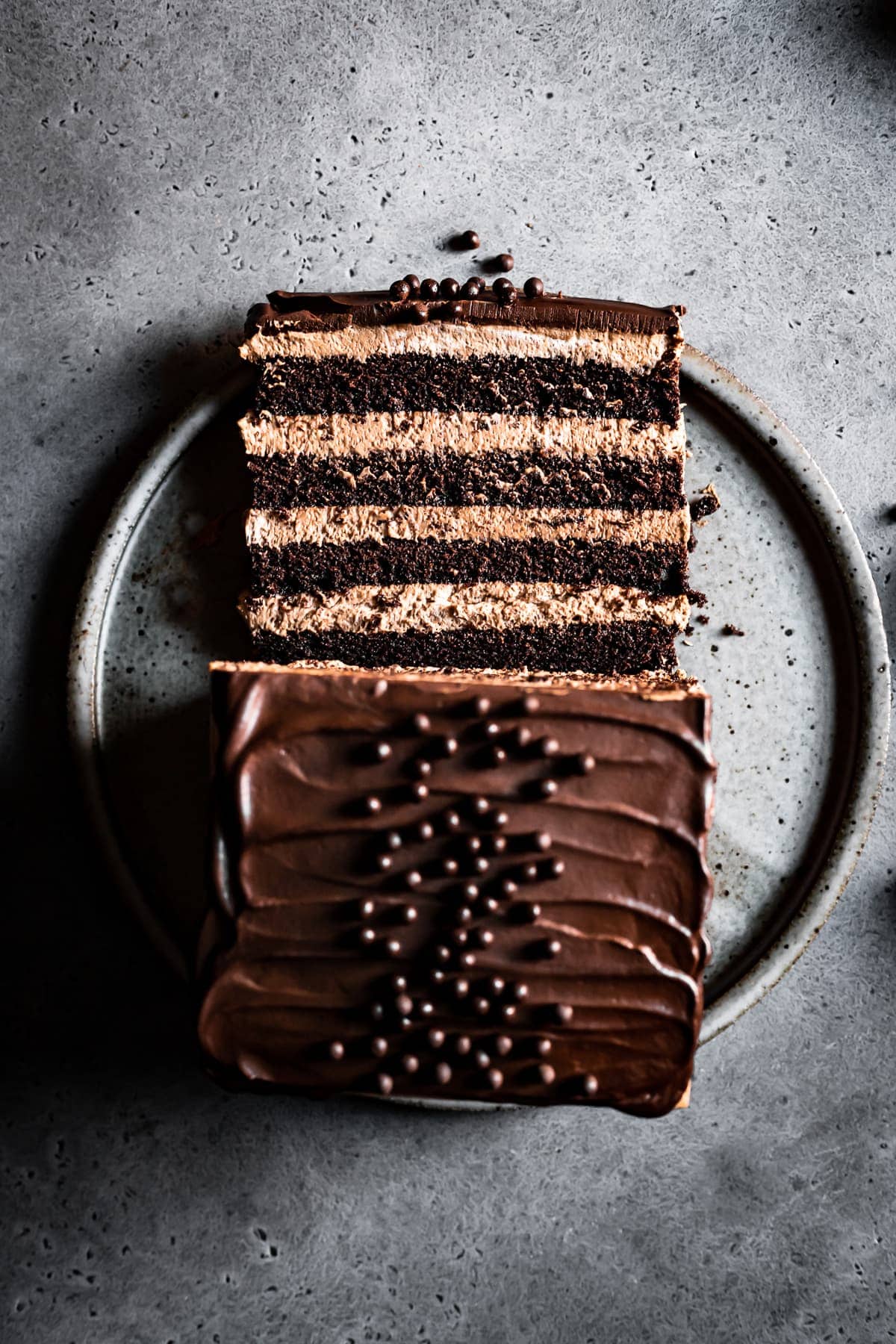 A moody image of a slice of chocolate mousse cake resting on its side on a grey speckled ceramic serving platter next to the remainder of the cake. The platter is on a grey stone surface.