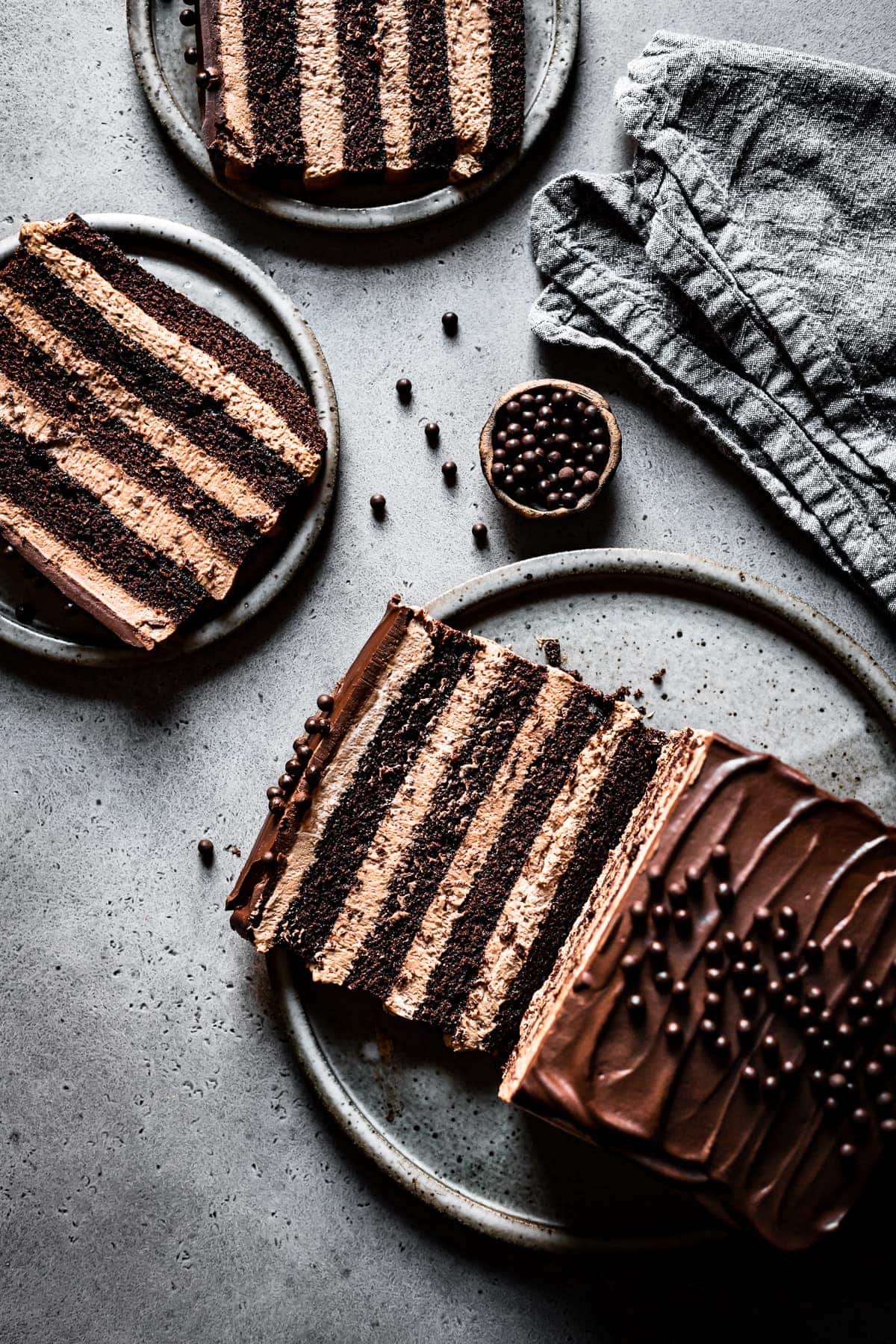 Three slices of brown layered cake on grey speckled ceramic plates on a grey speckled stone surface. A small bowl of edible chocolate pearls is nearby, along with a textured grey linen napkin. One of the cake slices is resting on the serving platter next to the remainder of the cake. 