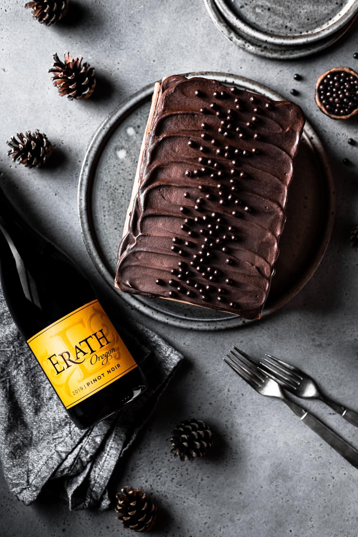 A rectangular chocolate cake topped with chocolate ganache on a circular grey speckled serving platter and a grey stone surface. A small bowl of edible chocolate pearls is nearby along with two grey ceramic plates, some pine cones and two forks. A bottle of red wine with an yellow orange label is lying to the left on a textured grey linen napkin