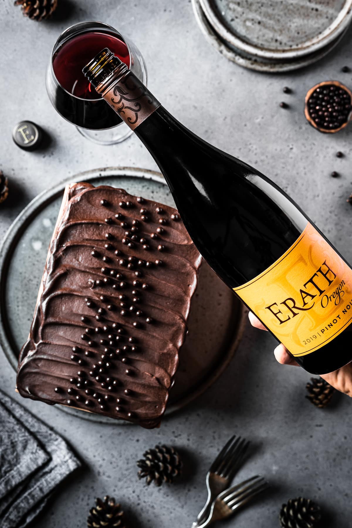 A rectangular chocolate cake topped with chocolate ganache on a circular grey speckled serving platter and a grey stone surface. A small bowl of edible chocolate pearls is nearby along with two grey ceramic plates, some pine cones and two forks. A hand reaches into the scene to pour a bottle of red wine with an yellow orange label into a wine glass. 