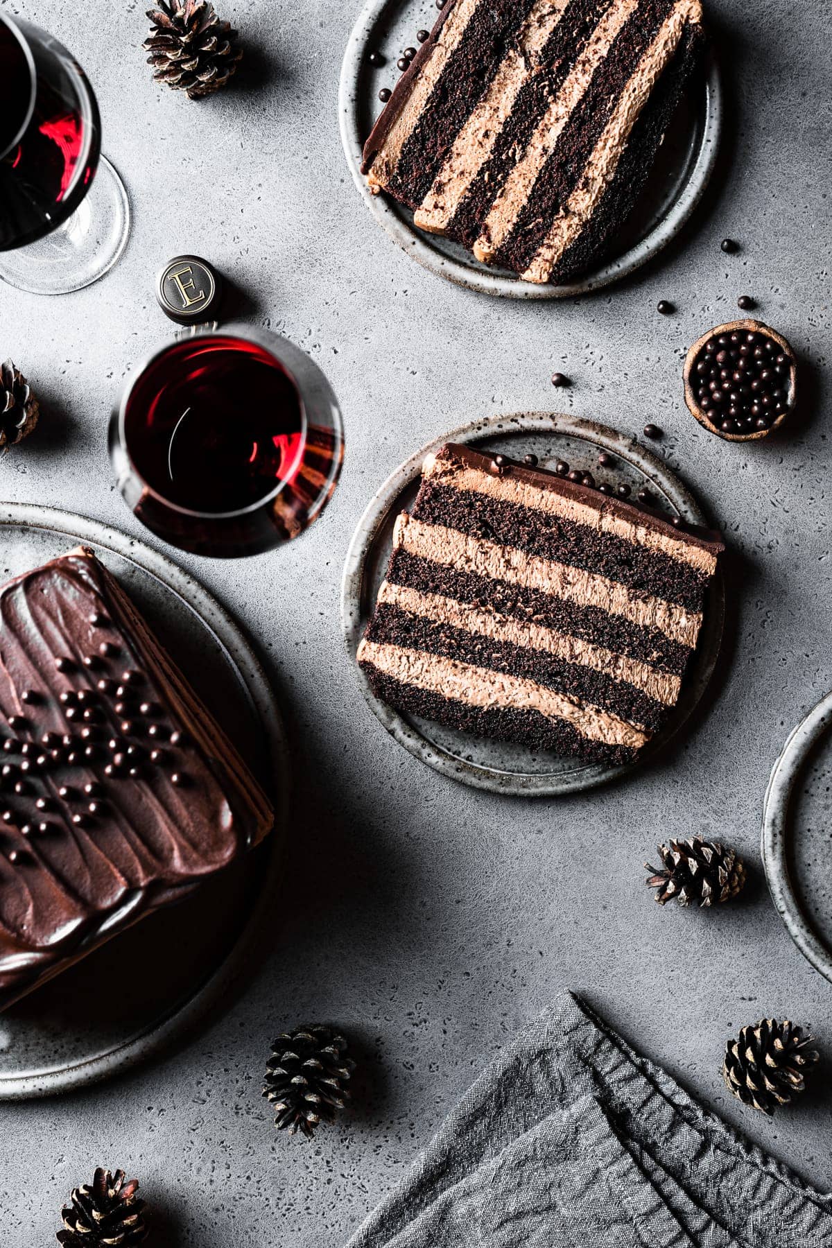 Two slices of chocolate cake on grey speckled ceramic plates on a grey speckled stone surface. A small bowl of edible chocolate pearls is nearby, along with a textured grey linen napkin, two glasses of red wine, and pine cones. A serving platter nearby holds the remainder of the cake. 