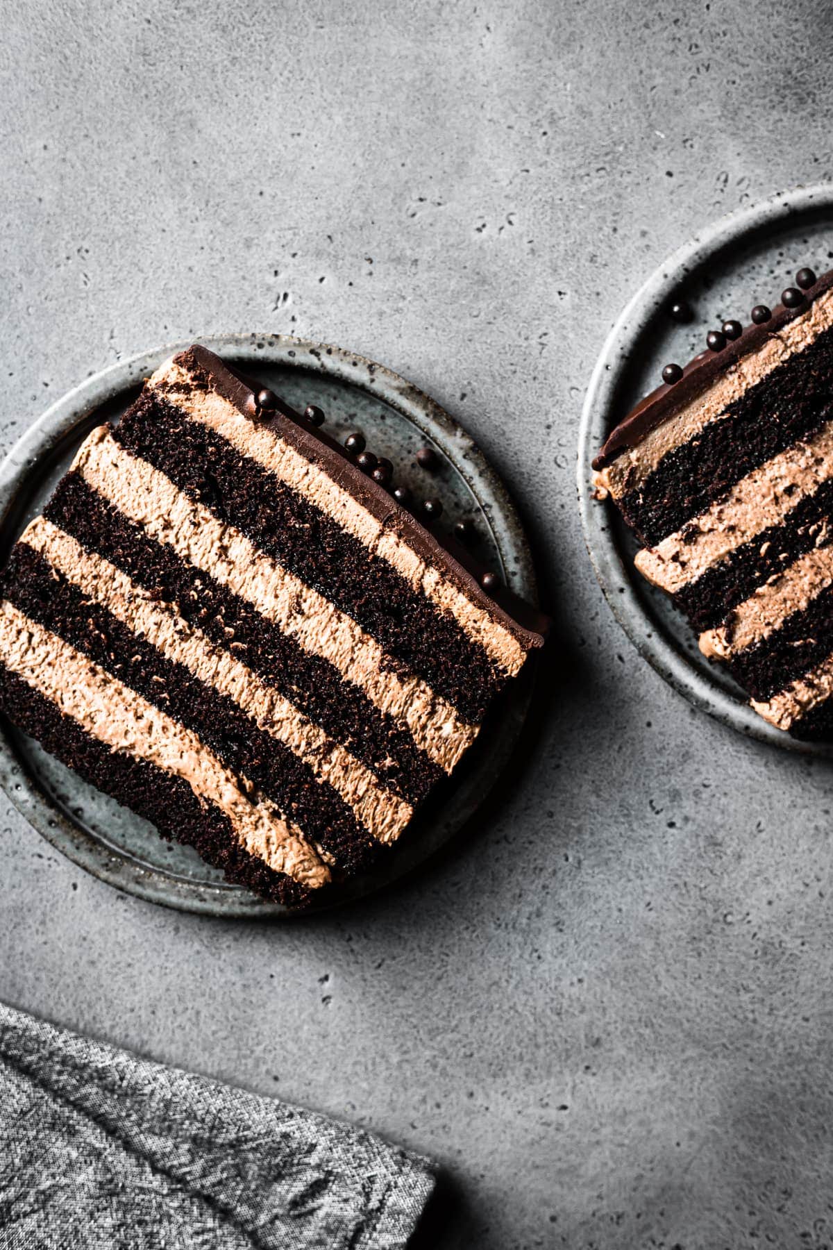 A close up of two rectangular slices of layered chocolate cake on grey speckled ceramic plates on a grey speckled stone surface. 