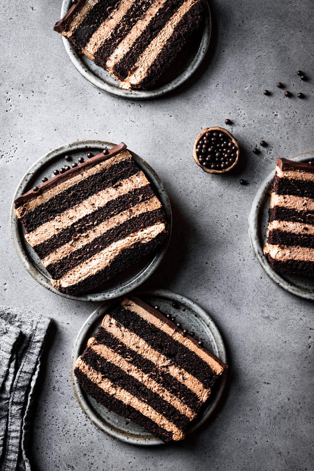 Four slices of chocolate cake on grey speckled ceramic plates on a grey speckled stone surface. A small bowl of edible chocolate pearls is nearby, along with a textured grey linen napkin. 