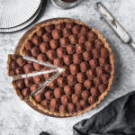 Two slices are cut out of a chocolate tart. It rests on a round wooden cutting board on a grey stone surface. There are plates, a knife and a napkin nearby.