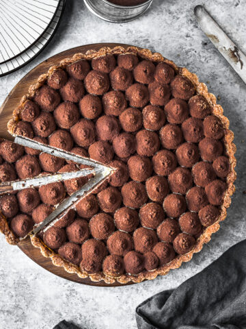 Two slices are cut out of a chocolate tart. It rests on a round wooden cutting board on a grey stone surface. There are plates, a knife and a napkin nearby.