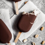 Chocolate dipped ice cream bars on parchment paper squares on a grey stone surface. One bar has bites out of it revealing the light brown interior. Peanuts are sprinkled nearby.