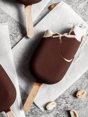 Chocolate dipped ice cream bars on parchment paper squares on a grey stone surface. One bar has bites out of it revealing the light brown interior. Peanuts are sprinkled nearby.