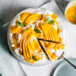 A coconut cake with baked meringue is decorated with vibrant yellow mango slices and mango puree. It rests on a white parchment square on a marble surface. A slice is being lifted out with a cake server.