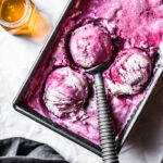 Three scoops of vibrant purple ice cream in a metal container full of ice cream. A vintage metal ice cream scoop holds one of the balls. A small pot of honey glows with the light shining through it at upper left. The background is white parchment paper.