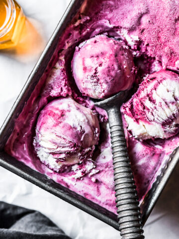 Three scoops of vibrant purple ice cream in a metal container full of ice cream. A vintage metal ice cream scoop holds one of the balls. A small pot of honey glows with the light shining through it at upper left. The background is white parchment paper.