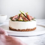 A small cheesecake on a white platter, topped with figs and rosemary, resting on a white marble surface. There is a pink napkin out of focus in the foreground.
