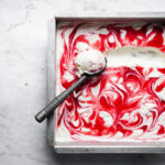 A container of ice cream with a dramatic red swirl of fruit on the top. A vintage metal ice cream scoop has left a trail through the ice cream and holds a full scoop as it rests atop the container. The entire container rests on a marble surface.