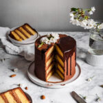 A golden layer cake with dark chocolate buttercream sits on a white ceramic plate with two slices cut out of the cake, revealing the layers. The cake rests on a white marble surface with the two slices on plates nearby.