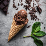 A waffle cone of chocolate ice cream rests on a grey stone surface. It is surrounded by a bar of chocolate, chopped chocolate shards, and a sprig of mint.