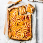A golden brown rectangular peach galette rests atop whipte parchment paper on a wooden cutting board. Two squares are cut out of the top right corner with a wooden handled paring knife.