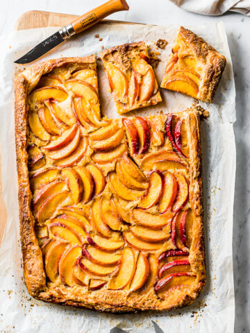 A golden brown rectangular peach galette rests atop whipte parchment paper on a wooden cutting board. Two squares are cut out of the top right corner with a wooden handled paring knife.