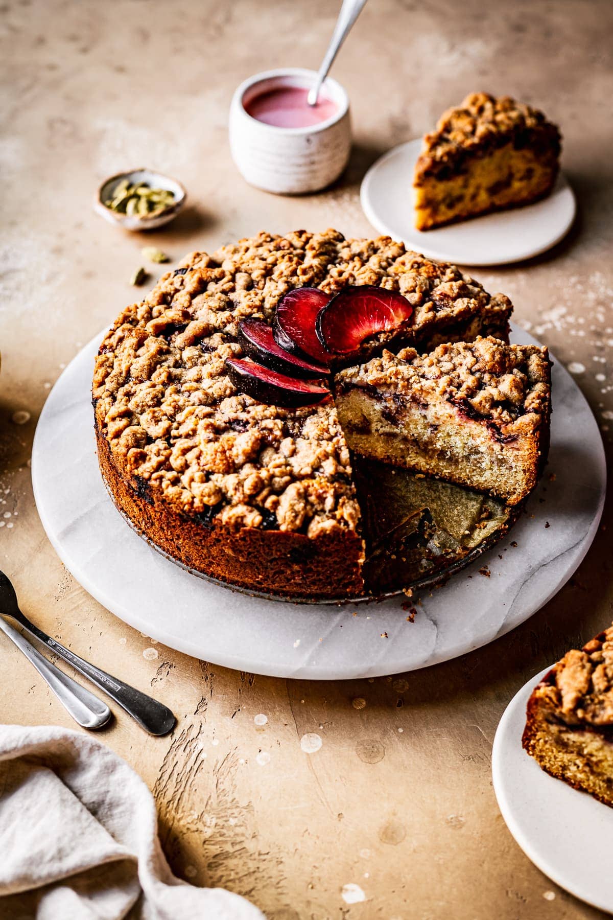 A round cake with several slices cut out of it on a marble platter on a warm tan surface. There is a fan of sliced plums on top of the cake. Nearby are several slices on small plates, a container of pink glaze, several forks, a natural linen napkin, and a small bowl of green cardamom pods.