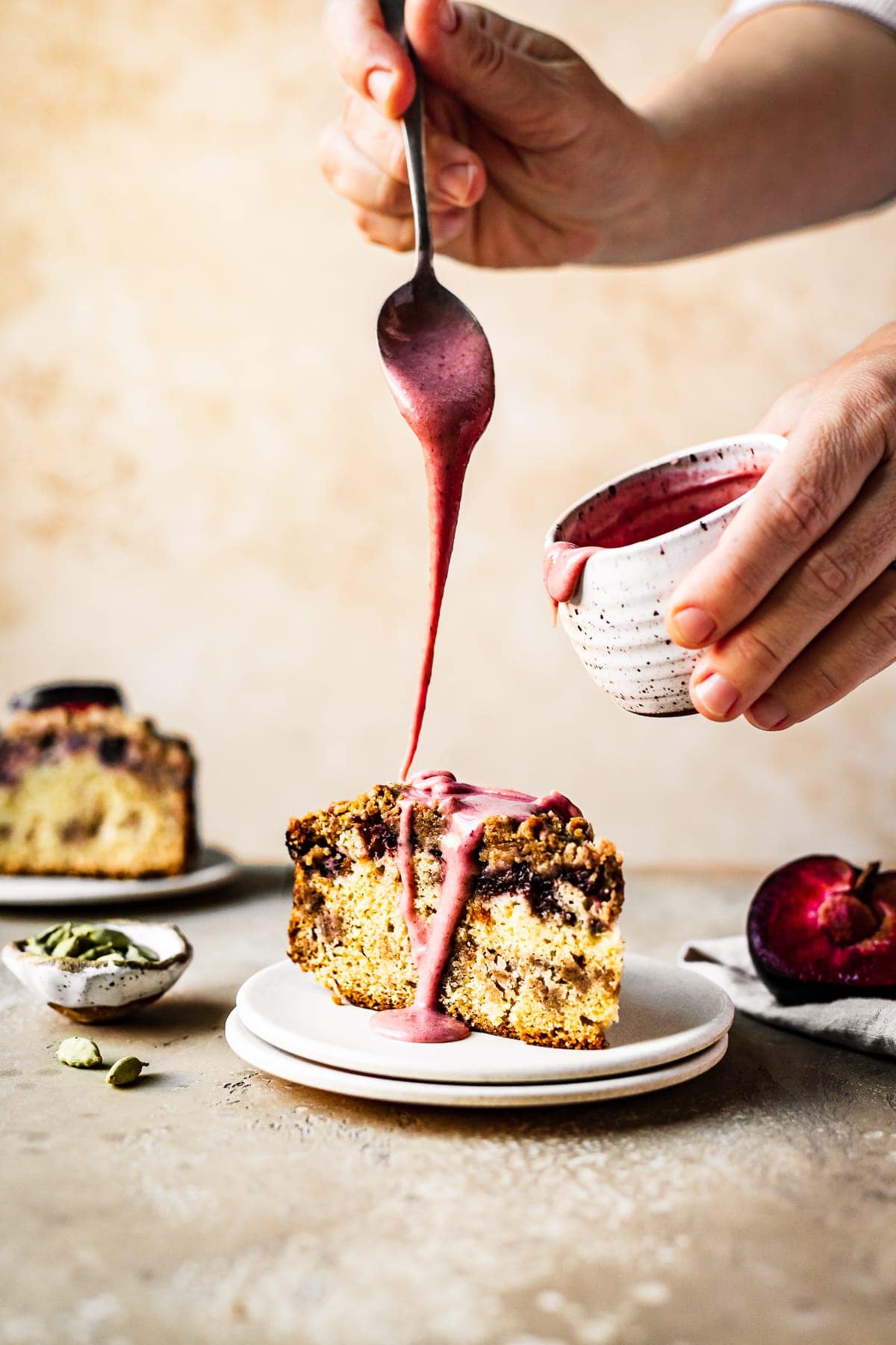 Hands reach into the frame drizzling a spoonful of pink glaze onto a slice of coffee cake. The other hand holds a white speckled ceramic jar of glaze. The surface and background are both a warm tan color. Resting next to the cake slice is half a plum, a small bowl of cardamom pods, and a slice of cake in the background.