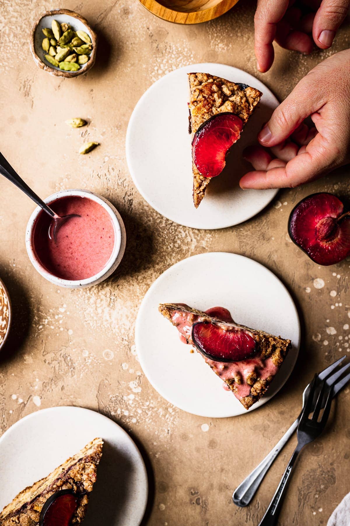 Hands reach into the frame towards slices of cake on light colored ceramic plates topped with plum slices. The background is a warm tan color. Surrounding the cake slices are forks, sliced plum halves, a bowl of pink glaze, and a small bowl of green cardamom pods.