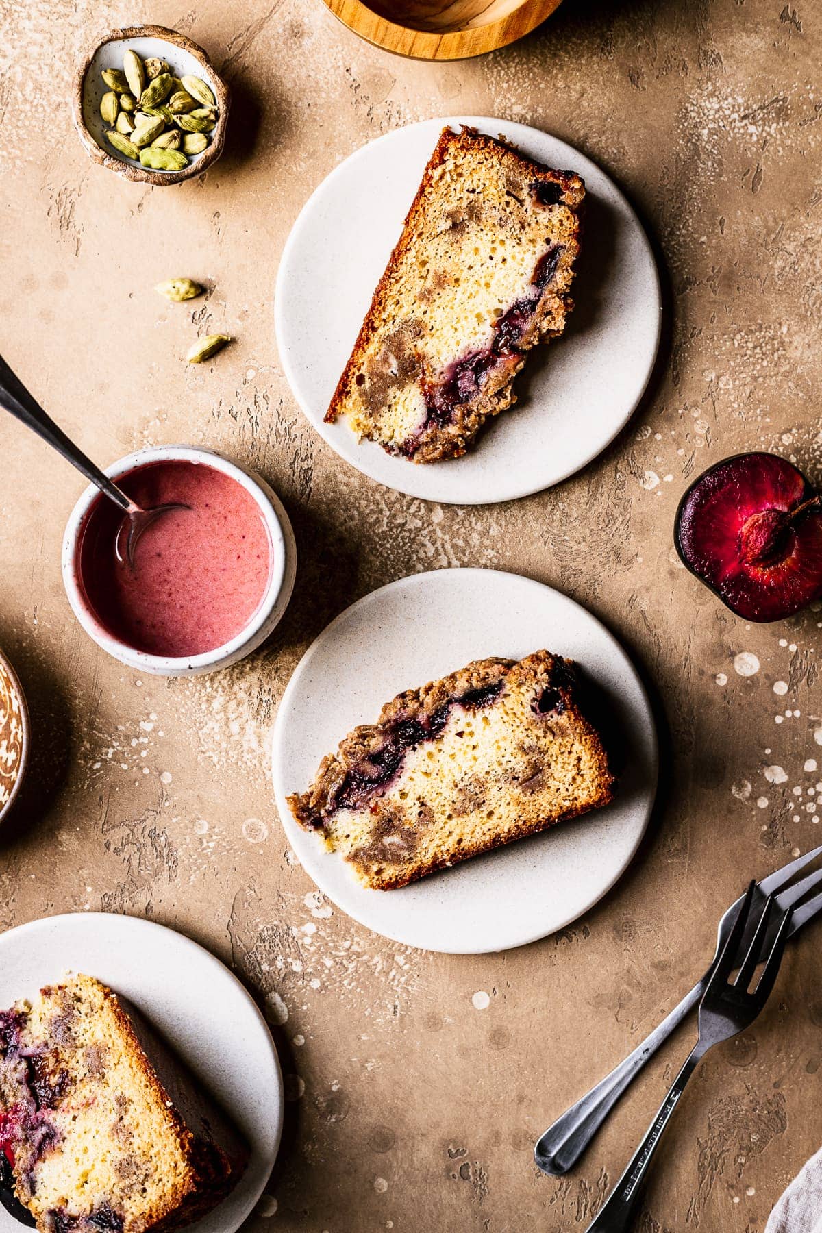 Slices of cake rest on their sides on light colored ceramic plates topped with plum slices. The background is a warm tan color. Surrounding the cake slices are forks, sliced plum halves, a bowl of pink glaze, and a small bowl of green cardamom pods.