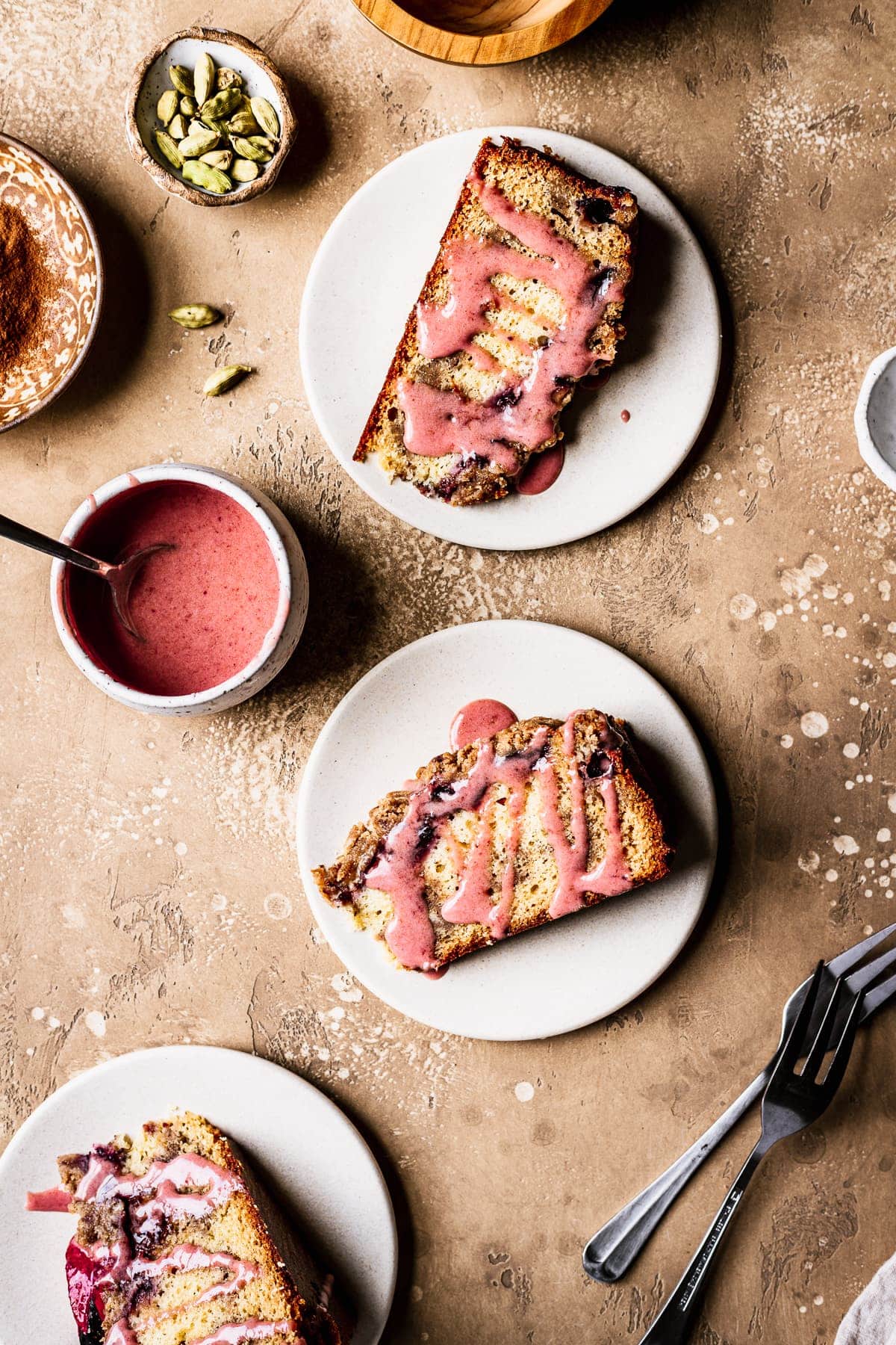 Slices of cake drizzled with pink glaze rest on their sides on light colored ceramic plates topped with plum slices. The background is a warm tan color. Surrounding the cake slices are forks, sliced plum halves, a bowl of pink glaze, and a small bowl of green cardamom pods.